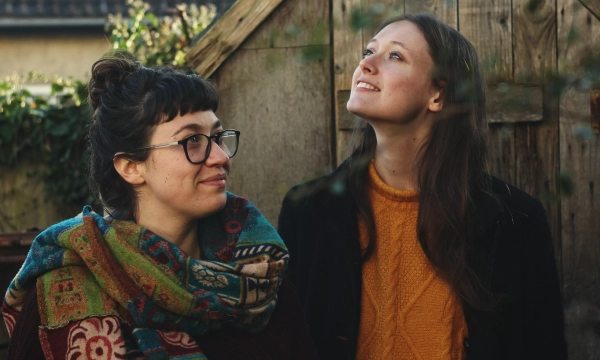 Two women stand outdoors, smiling and looking upward, surrounded by greenery and a wooden backdrop. One wears a colorful scarf and glasses, while the other has long hair and a cozy orange sweater.
