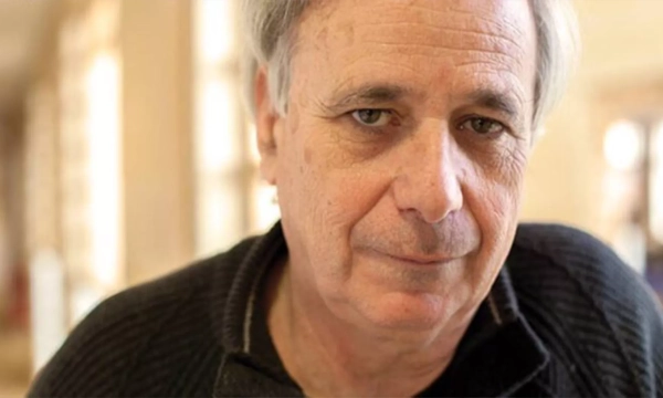 A middle-aged man with gray hair, wearing a black sweater, gazes directly at the camera in a softly lit interior setting.