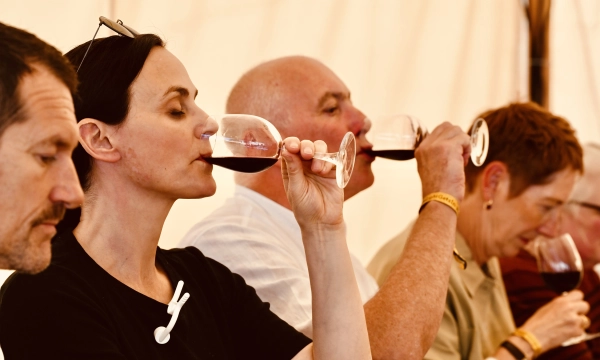 A group of people seated at a table, savoring red wine from glasses inside a tent. The focus is on a woman in a black shirt, enjoying her drink.