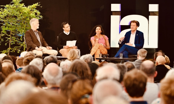 A panel discussion featuring four speakers, with a diverse audience in view. The setting includes a plant and a large illuminated sign in the background.