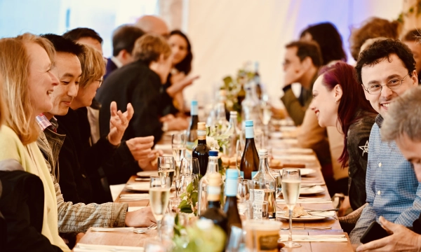 A long dinner table filled with people engaged in conversation, surrounded by bottles of wine and glasses, set in a festive atmosphere.