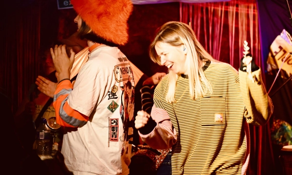 A lively indoor scene featuring a person in a colorful hat dancing joyfully, alongside another person wearing a striped shirt, with a vibrant background of lights and decorations.