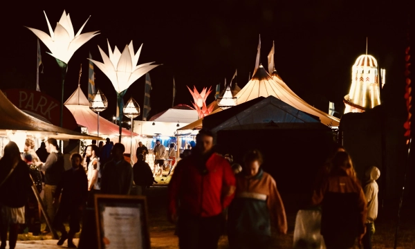 A vibrant night scene of a festival with illuminated flower-shaped decorations, tents, and people walking through the grounds, creating a lively atmosphere.