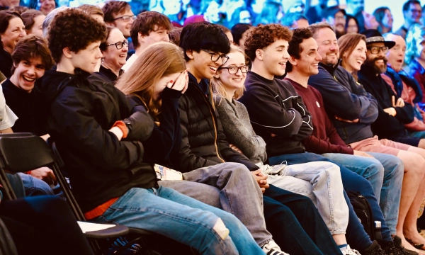 A diverse audience of young people and adults enjoying a lively performance, smiling and laughing in a brightly lit setting.