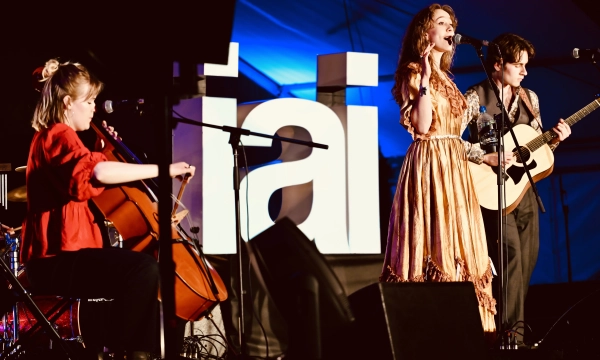 A singer in a flowing dress performs on stage with two musicians: one playing cello and the other holding an acoustic guitar, against a backdrop of the word "tai."