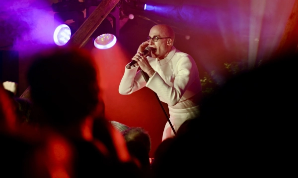 A performer in a white outfit passionately sings into a microphone on stage, surrounded by colorful lighting and an audience in shadow.