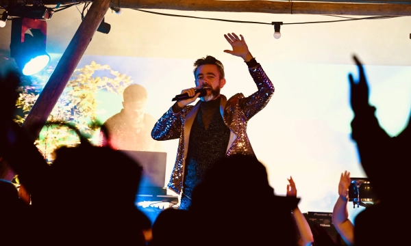 A performer in a sparkly jacket sings passionately on stage, engaging a lively audience with raised hands, colorful lighting, and an enthusiastic atmosphere.
