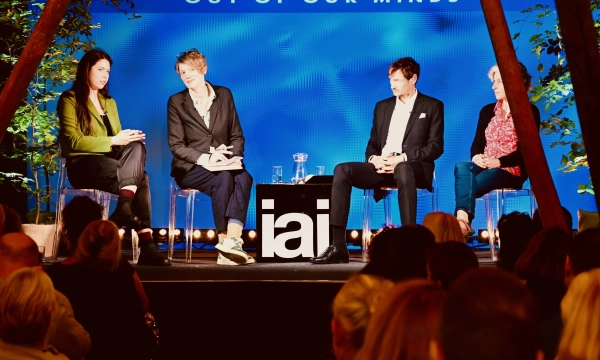 A panel discussion with four speakers seated on stage, addressing an audience, with a backdrop reading "OUT OF OUR MINDS" and the logo "iai."
