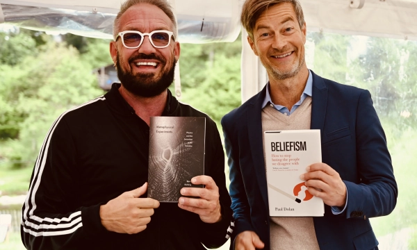 Two men holding books, one wearing a black tracksuit with sunglasses and the other in a blue blazer, both smiling at the camera.