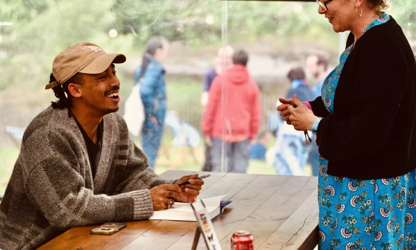 A man in a cozy sweater smiles while sitting at a table, talking to a woman in a blue dress, with people in the background.