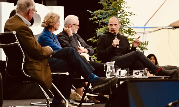 A panel discussion featuring four individuals seated, including a person speaking into a microphone, with a backdrop of greenery and a tent setting.