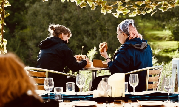 Two individuals sitting at a table outdoors, enjoying sandwiches with a scenic backdrop of greenery and nature.