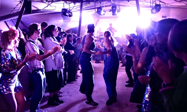 A lively dance floor scene inside a tent, featuring people in colorful outfits cheering and clapping as a couple dances under vibrant lighting.