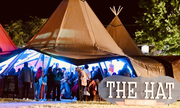 A lively outdoor bar and music venue featuring large teepee-like structures, with a crowd enjoying the evening atmosphere illuminated by colorful lights.