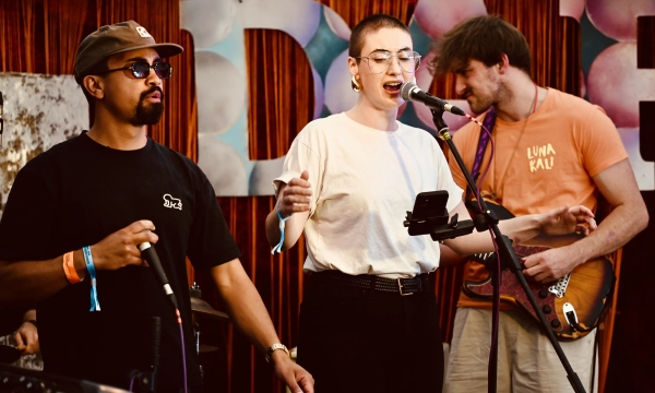 A musical performance featuring a male artist rapping, a female vocalist singing, and a male musician playing guitar, with a vibrant backdrop and stage setup.