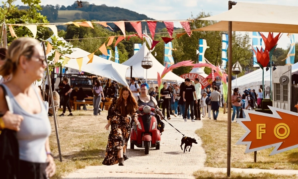 A lively outdoor festival scene with colorful flags, tents, and people walking, including a woman pushing a scooter and someone walking a dog.