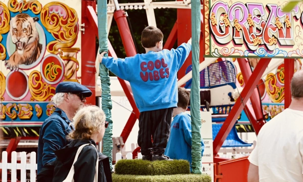 A child wearing a blue "COOL VIBES" sweatshirt stands on a green platform at a carnival, surrounded by adults and colorful fair attractions.