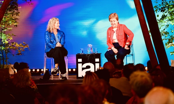 Two speakers sit on stage during an event, with a colorful backdrop and audience in front. One wears a blue jacket, the other a red one.