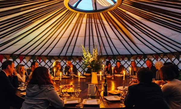 A group of people gathered around a large table inside a yurt, with candlelight illuminating the intimate setting and floral centerpiece.