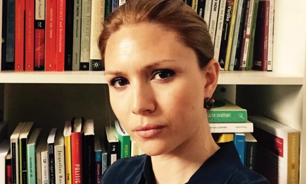 A woman with straight hair is taking a selfie in front of a bookshelf filled with various academic books.