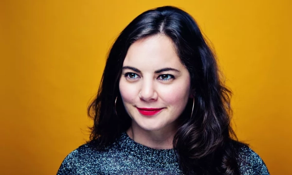 A smiling woman with dark hair and bright red lipstick against a vibrant yellow backdrop, wearing a sparkly black top.