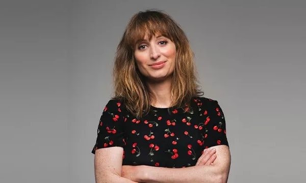 A woman with wavy brown hair and a black shirt featuring a cherry pattern stands confidently with her arms crossed against a gray background.