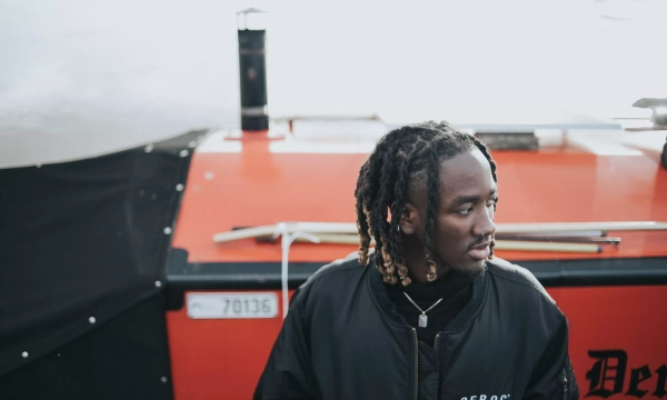 A young man with braided hair looking away from the camera, standing near a red boat with a black cover and the name "Devil" visible on it.
