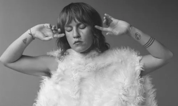 A person with shoulder-length hair and tattoos poses in a fluffy white garment, eyes closed, embodying a serene and introspective mood.