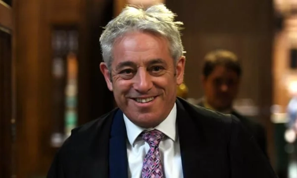A smiling man with white hair wearing a dark robe and tie, walking in a historic building, likely a governmental setting.