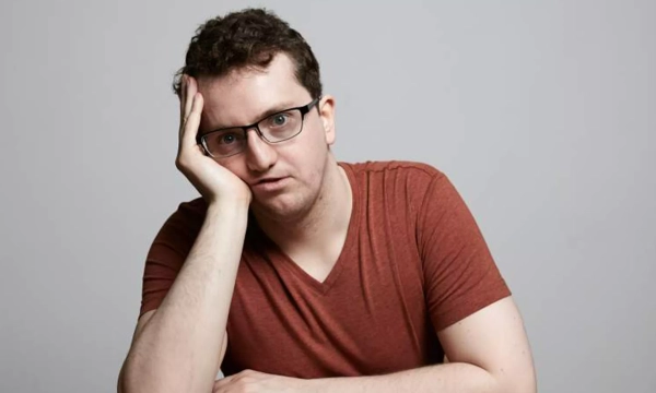 A man with glasses and curly hair rests his head on his hand, looking thoughtfully at the camera while wearing a casual red t-shirt against a plain background.