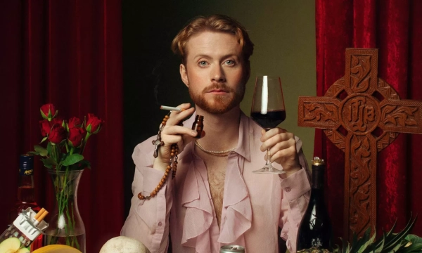 A person with a pink ruffled shirt holds a glass of wine and a smoking pipe, surrounded by flowers, food items, and a wooden cross in a theatrical setting.
