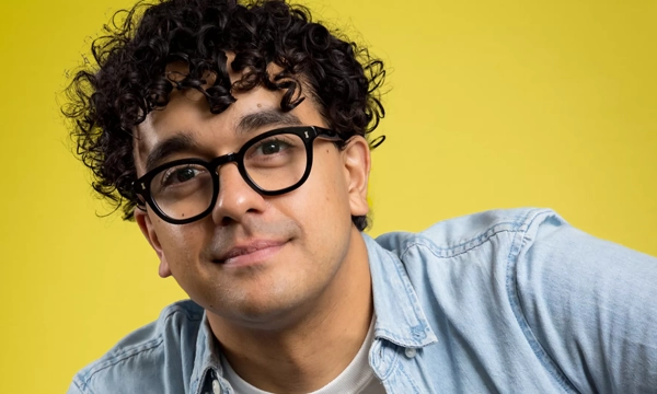 A young man with curly hair and glasses smiles against a vibrant yellow background, wearing a denim jacket over a t-shirt.
