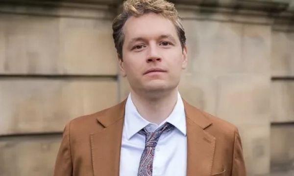 A well-dressed young man with light hair stands in front of a textured stone wall, wearing a brown blazer and patterned tie.