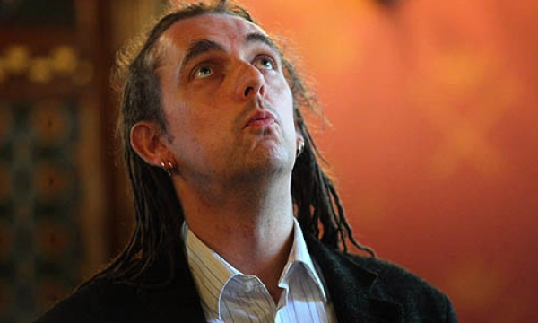 A man with dreadlocks gazes upward in a richly decorated room with warm-colored walls and intricate patterns.