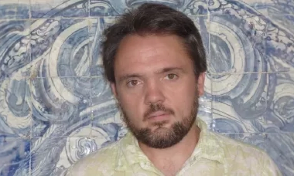 A man with a beard and short hair stands against a patterned blue and white tiled wall, wearing a light-colored shirt.