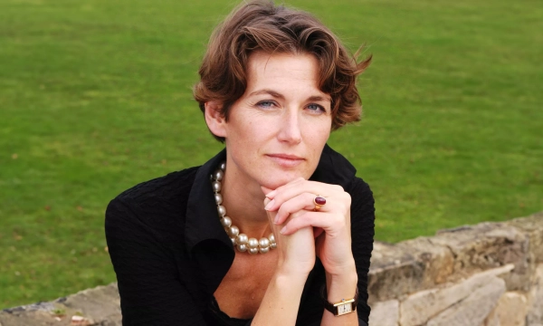 A woman in a black top with a pearl necklace resting her chin on her hands, sitting outdoors against a grassy background.