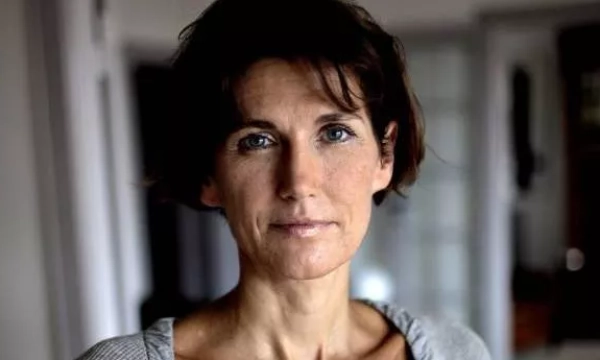 A middle-aged woman with short hair, wearing a gray sweater, gazes thoughtfully at the camera in a softly lit room.