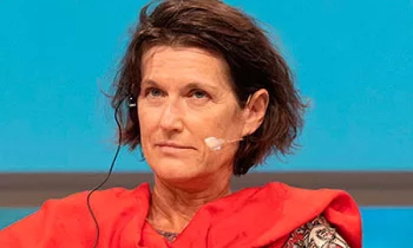 A woman with shoulder-length brown hair wearing a red shawl and a microphone headset, seated against a blue backdrop during a panel discussion.