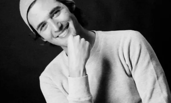 A smiling young man wearing a gray sweater and beanie, sitting casually with his hand on his chin against a dark background.