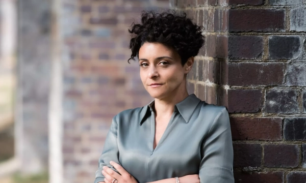 A woman with curly hair wearing a light green blouse stands against a brick wall, looking confidently at the camera with her arms crossed.