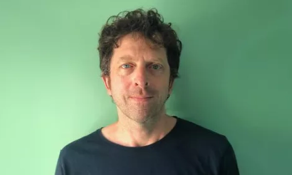 A man with curly hair and blue eyes stands against a green background, wearing a dark shirt and looking directly at the camera.