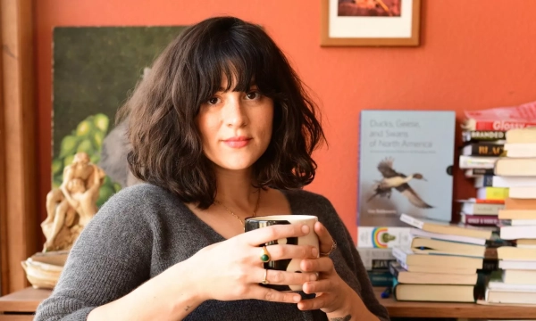 A woman with wavy, shoulder-length hair holds a black mug while seated in a cozy, book-filled room, featuring paintings and various books in the background.