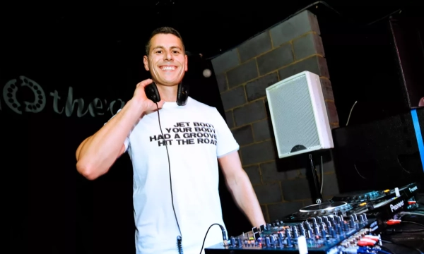 A DJ smiles while performing, wearing a t-shirt with a playful phrase, next to a sound mixer and speaker in a vibrant setting.