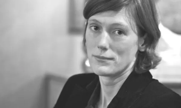 A woman with short brown hair and a serious expression, wearing a black jacket, sitting indoors with a blurred background.