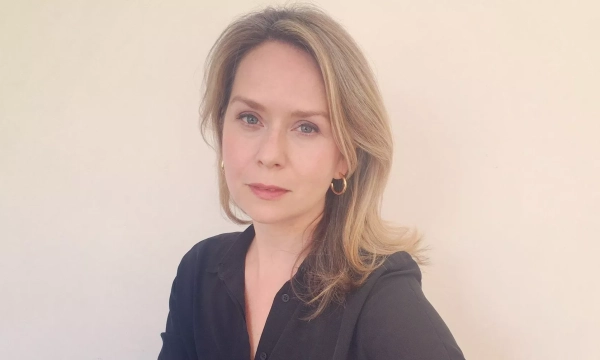 A woman with blonde hair and hoop earrings, dressed in a black shirt, stands against a neutral background, looking directly at the camera.