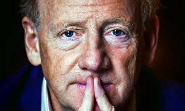 A close-up portrait of a mature man with light brown hair and blue eyes, thoughtfully resting his chin on his hands.