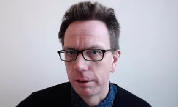 A man with glasses and styled hair looks directly at the camera, wearing a black sweater over a patterned shirt, against a plain background.