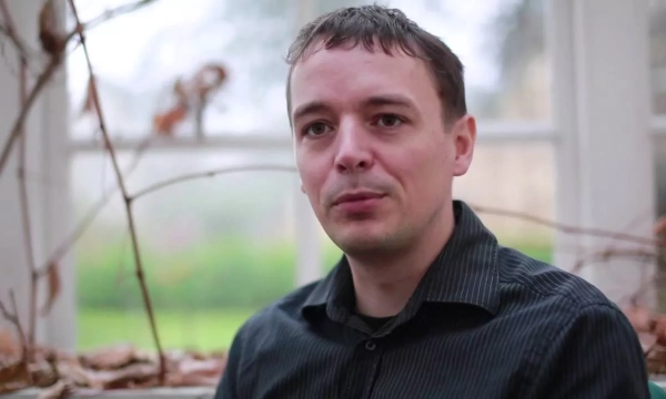 A man with short dark hair, wearing a black shirt, sits in a garden setting with blurred greenery in the background and dried plants in the foreground.