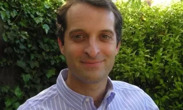 A man with brown hair, wearing a light blue striped shirt, smiles while sitting in front of green foliage in a sunny outdoor setting.
