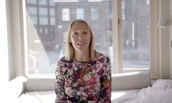 A woman with blonde hair sits on a bed in a bright room with large windows, wearing a floral patterned shirt, looking at the camera.